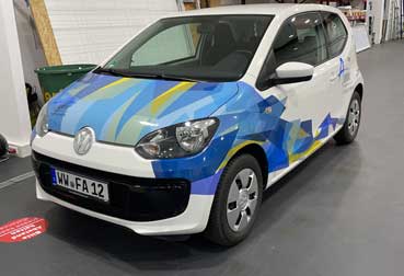 Volkswagen Up in weiß mit blau-gelben Dreiecken auf Haube und Seite foliert.